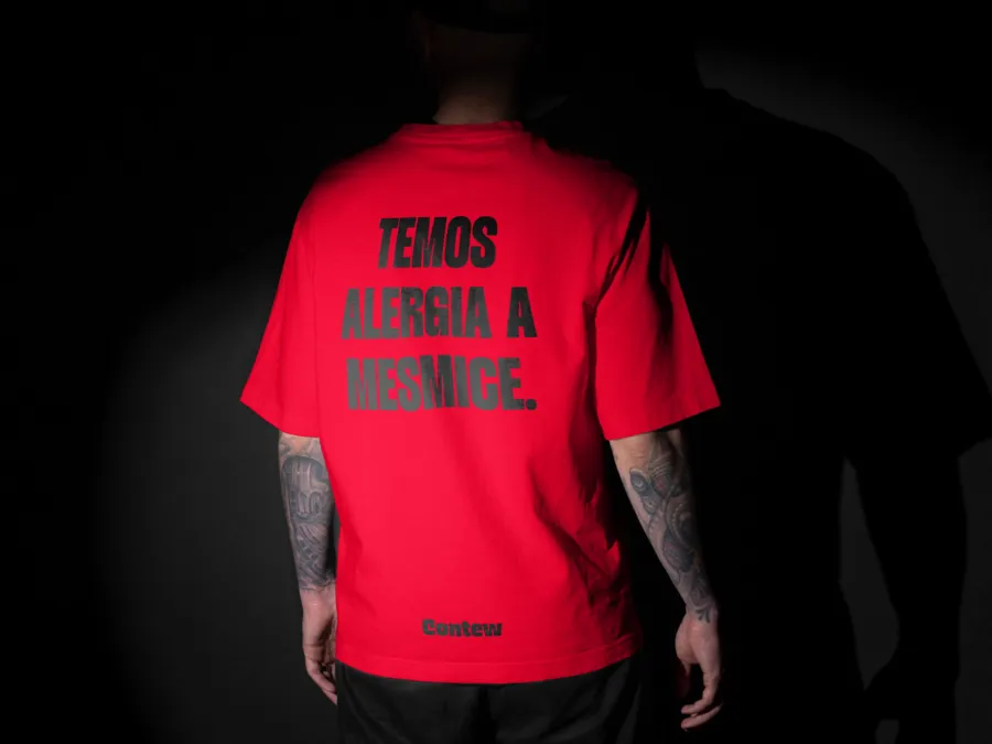Pessoa de costas vestindo camiseta vermelha com a frase 'Temos alergia a mesmice' estampada, em um ambiente escuro